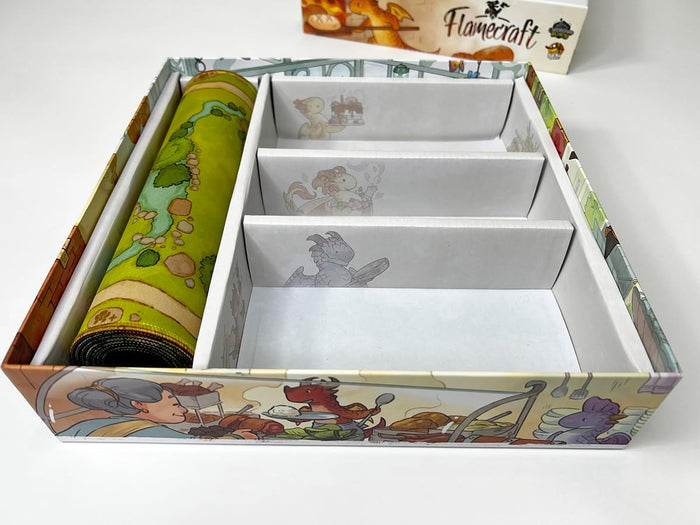 Open Flamecraft board game box with a rolled-up green playmat and illustrated cardboard organizer dividers.