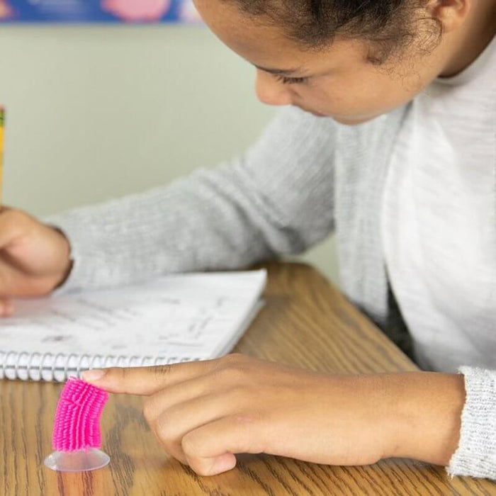 A child's finger flicks a small, pink, textured silicone stick with a suction cup base attached to a wooden desk.