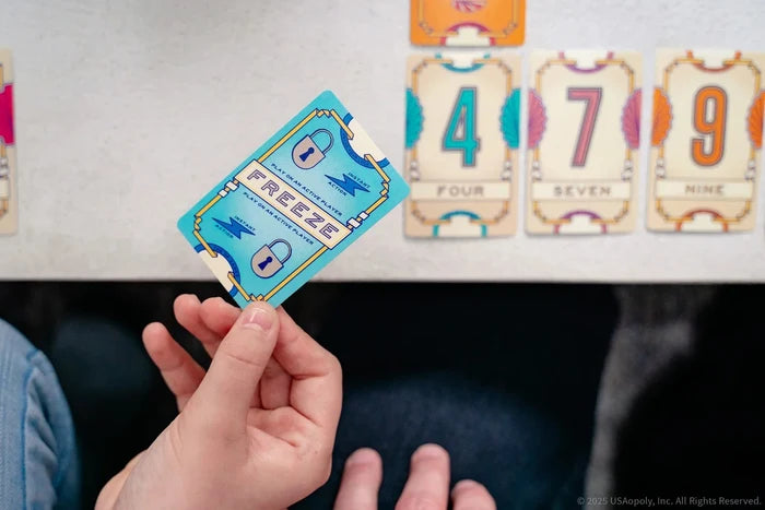 Hand holds a blue Freeze card with lock icons over numbered cards 4, 7, and 9 on a white table.