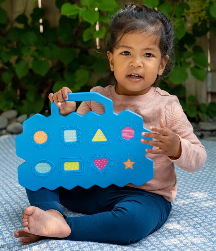 Toddler holding a blue activity board with a handle and colorful shapes like a heart, star, and triangle.