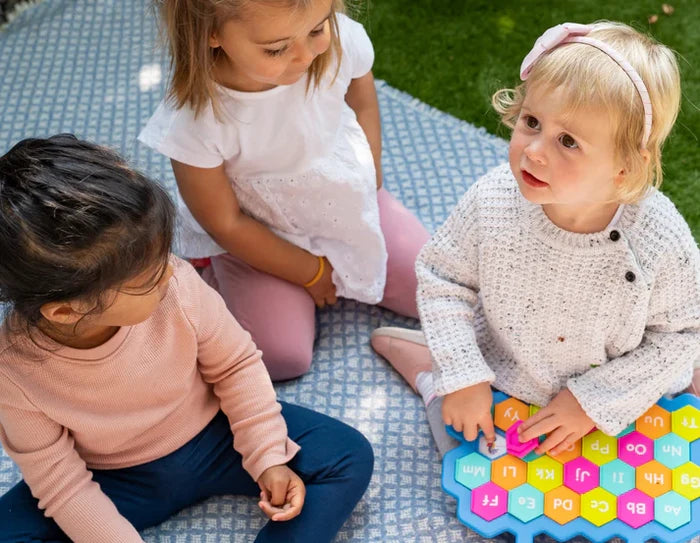 Three toddlers sit on a blanket playing with a blue hexagonal alphabet toy featuring colorful letter flaps.