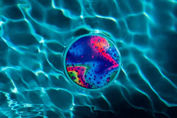 Colorful pink and blue patterned round disc floating on shimmering blue pool water with sunlight ripples.