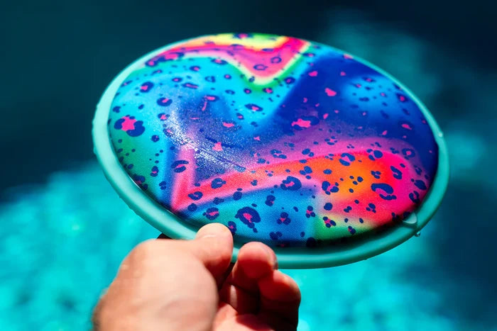 Hand holding a wet, colorful leopard-print flying disc with a light blue rim over a blurry turquoise pool.