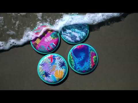 Four circular flying discs with bright tropical prints on wet sand as ocean water washes over them.