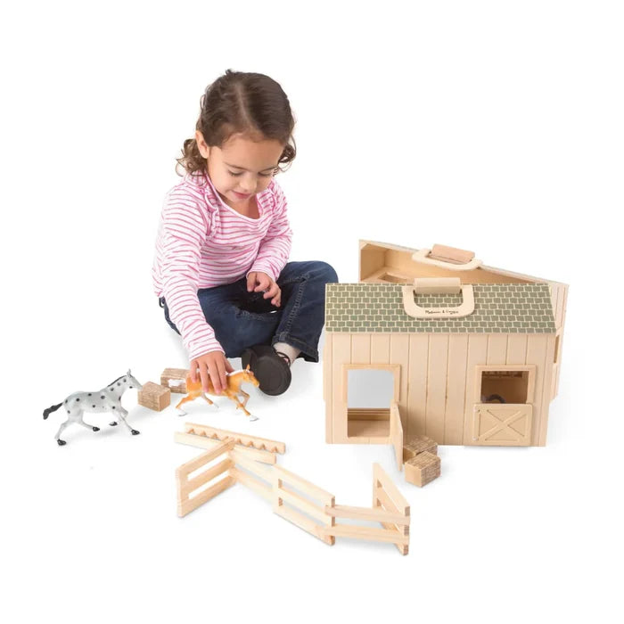 Child playing with a portable wooden toy stable featuring a green roof, two toy horses, fences, and hay bales.