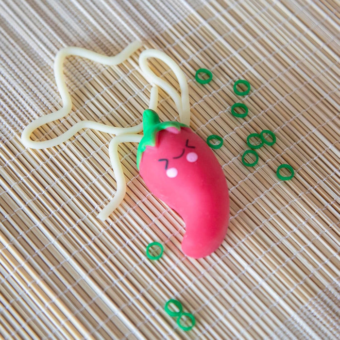 Red chili pepper toy with a cute face, stretchy white noodles, and small green rings on a bamboo mat.