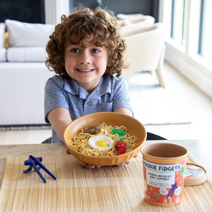 Child holding a bowl of toy ramen noodles with a smiley egg, beside blue chopsticks and branded product cup.