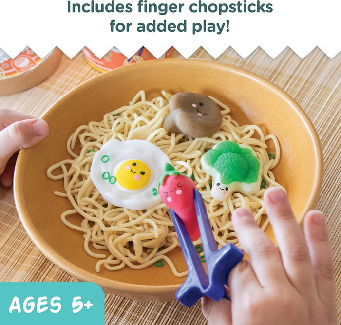 Child using purple finger chopsticks to pick up a red toy chili from a bowl of play ramen with smiley faces.