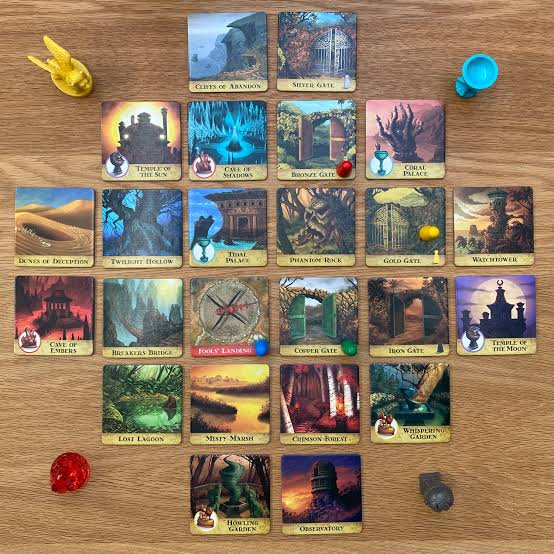 A board game layout featuring multiple illustrated cards arranged on a wooden surface, along with several colored tokens or pawns. The cards show fantasy-themed scenes and titles such as City of Ashram, Mithra Gate, Trials of the Sun, and others.