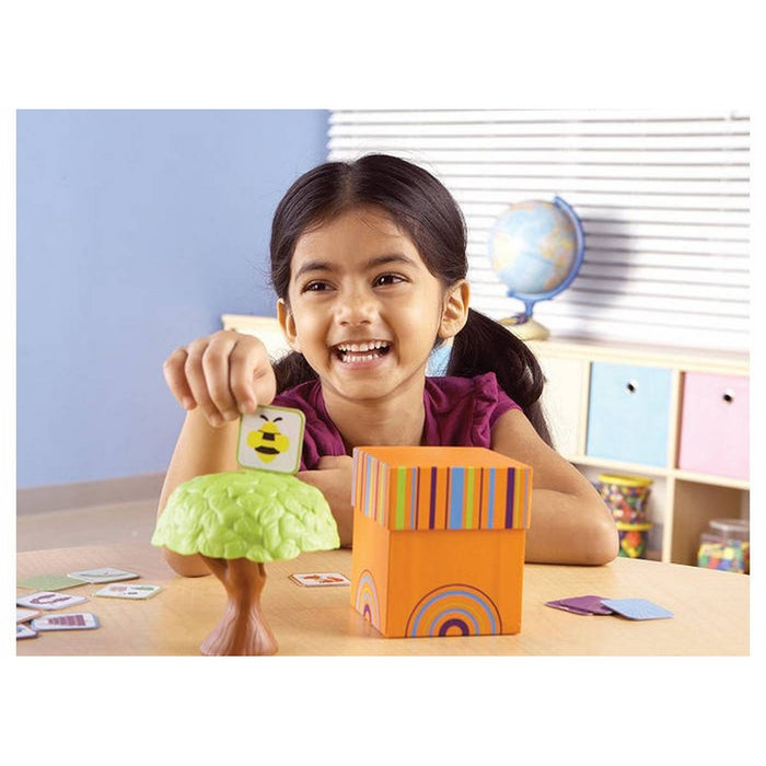 Smiling girl holds a bee card over a toy tree next to a colorful striped orange box during a learning game.