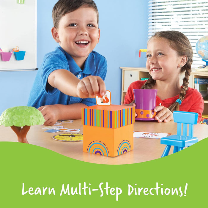 Two children play with an orange striped box, fox cards, a blue chair, purple cup, and green tree toy on a table.