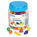 Clear jar with blue lid filled with colorful plastic fruit. Small apples, grapes, and bananas scattered in front.