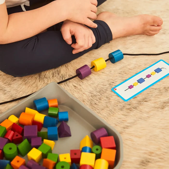Child stringing colorful wooden beads to match a pattern card, with a tray of assorted shapes on a plush rug.