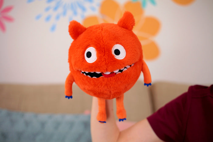 Round fuzzy orange monster puppet with wide white eyes and a toothy smile, shown being worn on a person's arm.