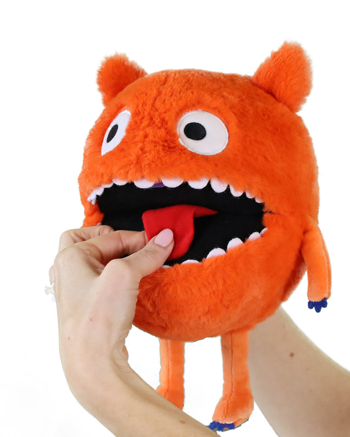 Round orange fuzzy monster puppet with wide eyes, white teeth, and a red tongue being touched by a hand.