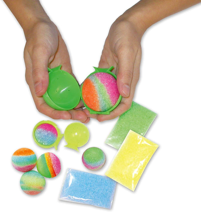Hands reveal a textured multi-color ball in a mold, with finished bouncy balls and packets of colored granules.