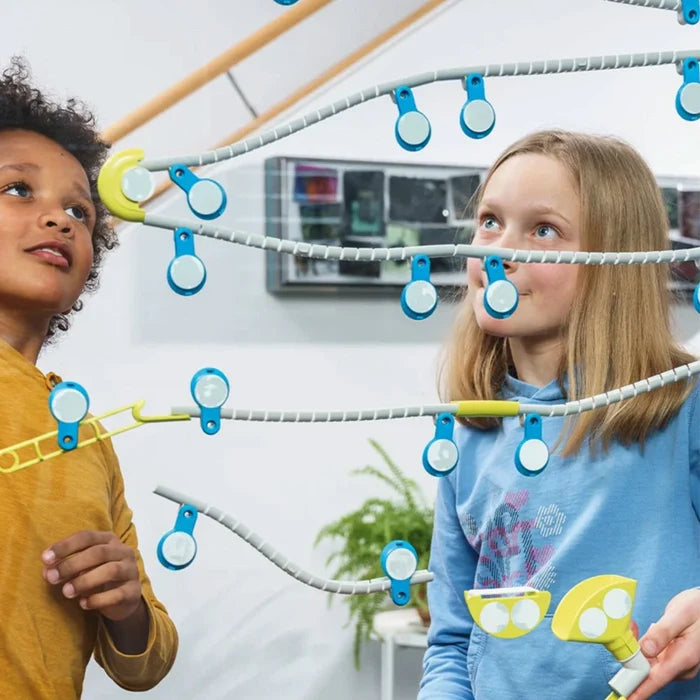 Two children look at a flexible gray marble run featuring blue circular clips and yellow curved plastic pieces.