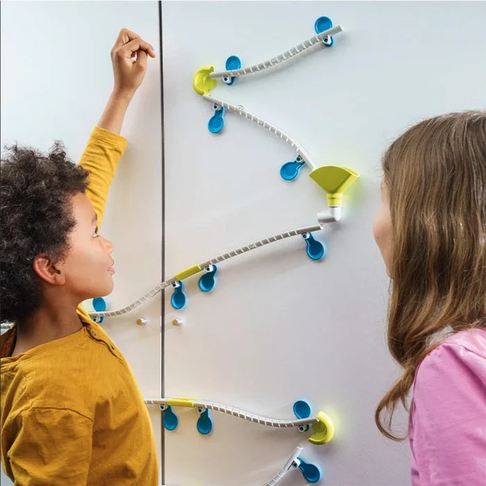 Children play with a green and white wall-mounted marble run attached with blue suction cups to a white surface.