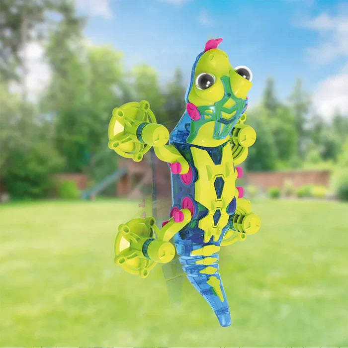Translucent blue and green robotic gecko with suction cup feet climbing a vertical glass surface in a backyard.