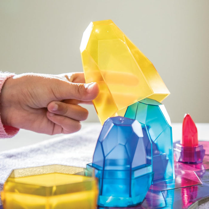 A child's hand stacks translucent yellow, teal, and blue geometric crystal toy blocks on a game board.