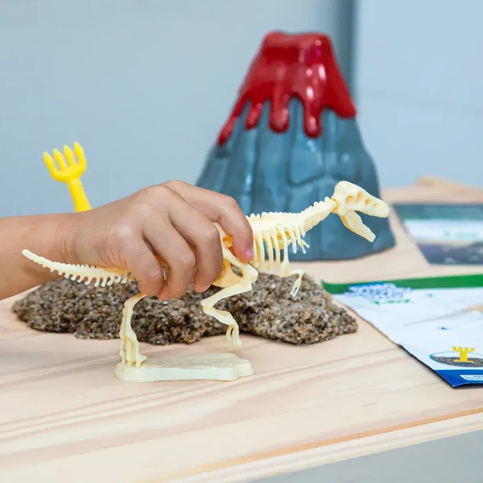 Child's hand holding a cream T-Rex skeleton toy next to a grey and red toy volcano and gravel on a wooden table.