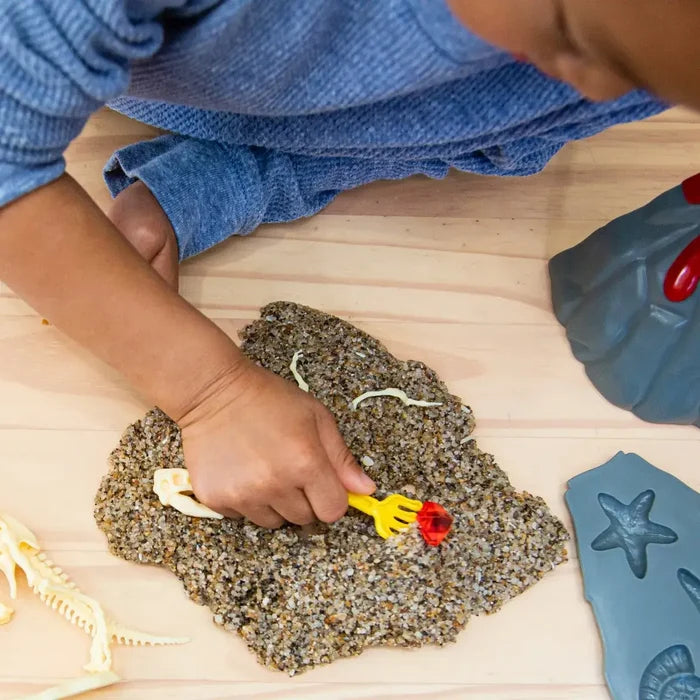 Child using a yellow tool to dig for dinosaur fossils and a red gem in textured play sand on a wooden surface.
