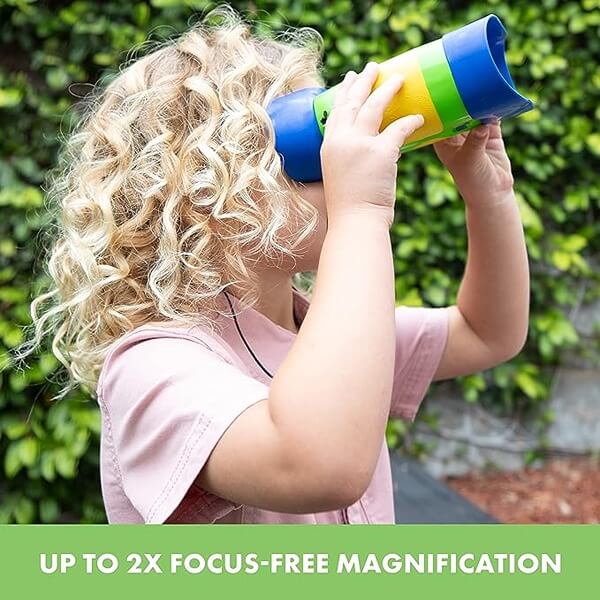 A curly-haired child looks through chunky blue and yellow toy binoculars against a background of green leaves.