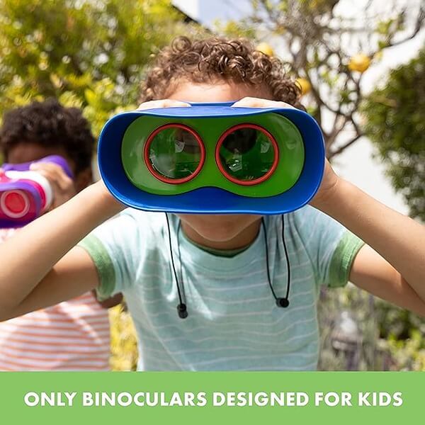 Child looking through blue and green toy binoculars with red-rimmed lenses in a sunny outdoor setting.