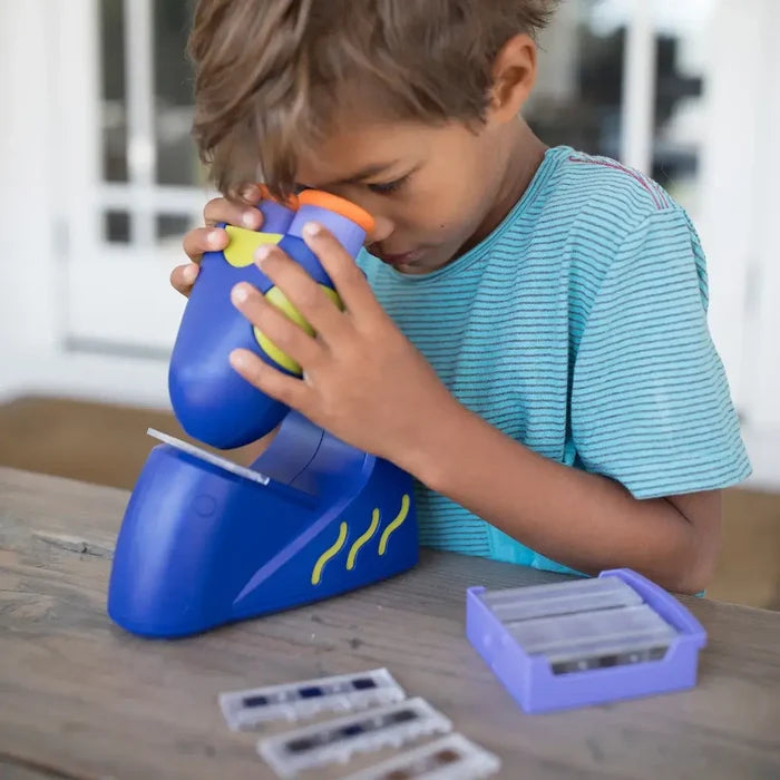 Child using a blue and green toy microscope with plastic slides and a purple storage box on a wooden table.