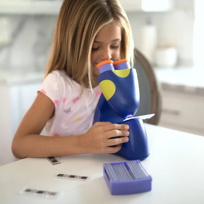 Young girl using a blue and yellow toy microscope with slides and a purple storage box on a white table.