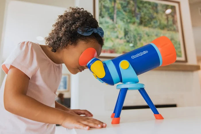 Young girl looks through a colorful blue and orange toy telescope on a tripod.