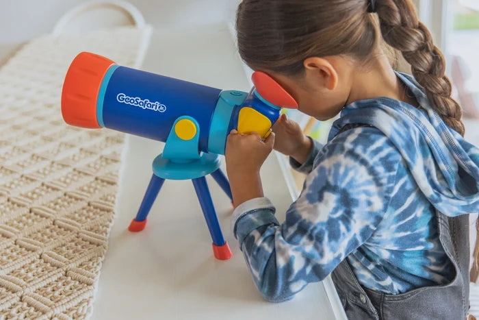 Young girl looking through a blue and orange GeoSafari Jr. toy telescope on a tripod with yellow accents.