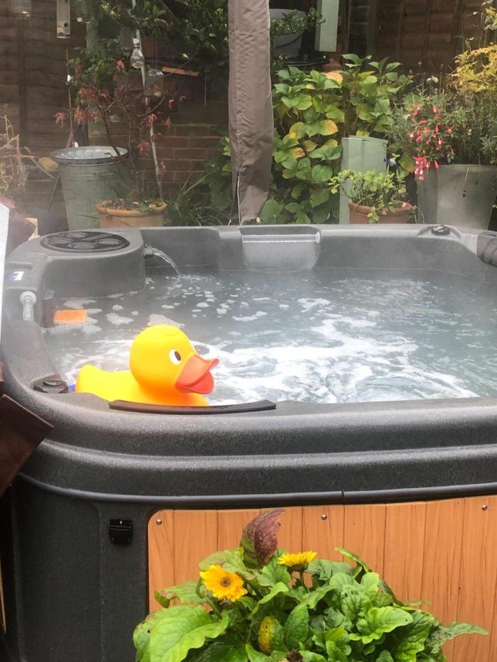 Giant bright yellow rubber duck with an orange beak floating in a bubbling grey outdoor hot tub.