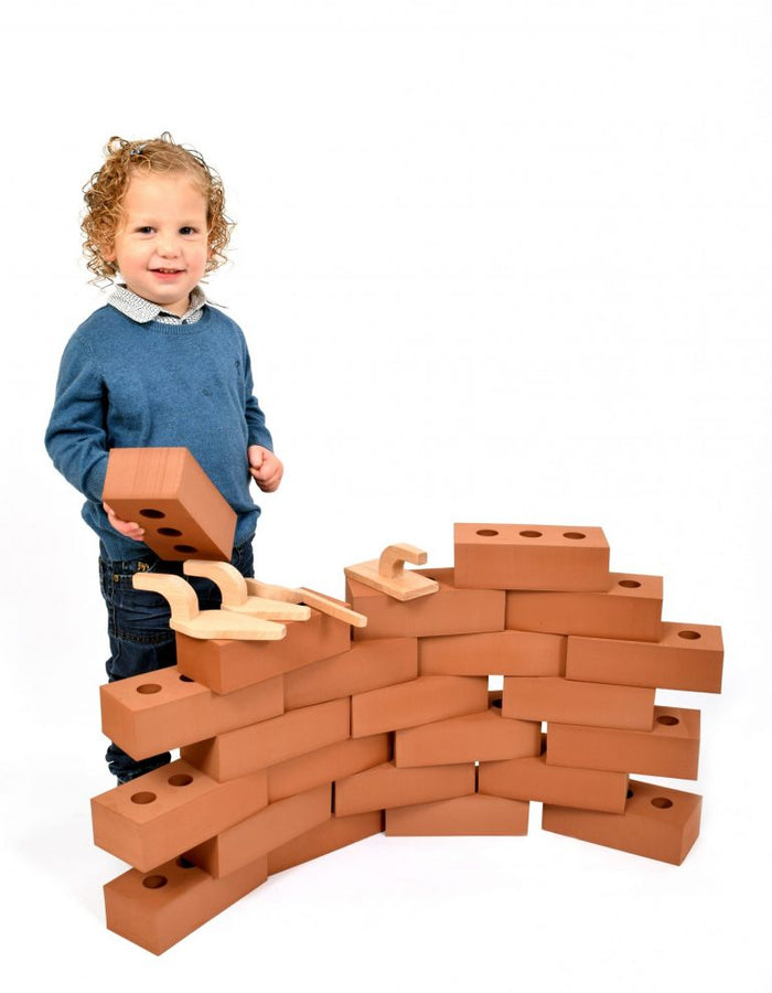 Child building a wall with large terracotta-colored foam bricks and wooden toy trowels.