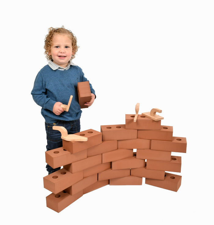 A toddler builds a curved wall using large, reddish-brown foam bricks with two holes and wooden toy tools.