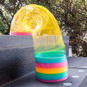 Giant translucent rainbow coil toy walking down dark gray stairs, featuring vibrant pink, orange, and blue layers.