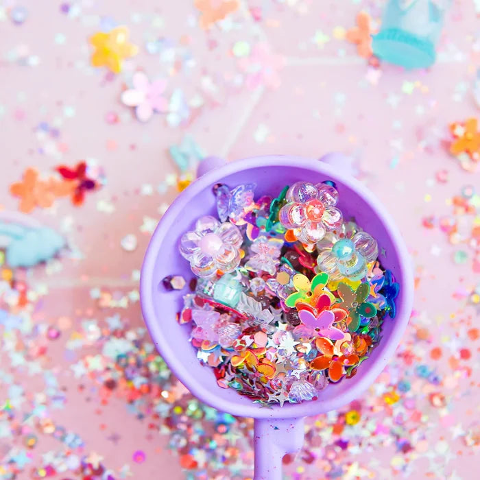 Colorful plastic flower, star, and butterfly charms filling a purple cup against a sparkly pink background.