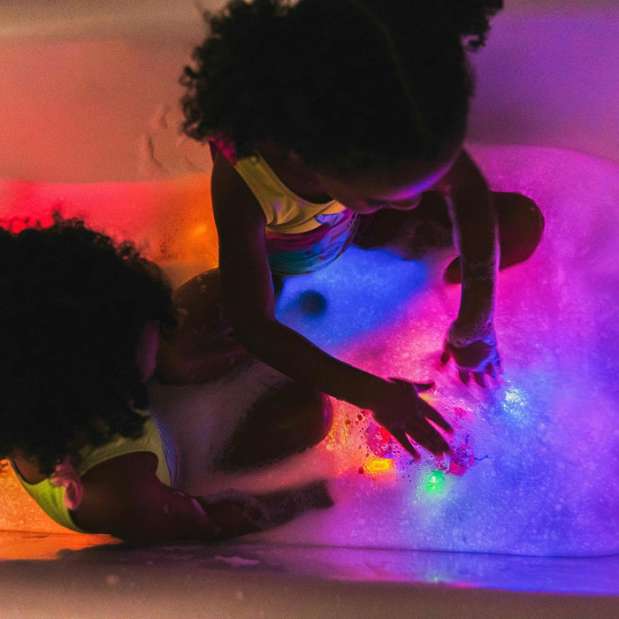 Two children in a bubble bath playing with colorful red, blue, and green light-up cubes that glow under the water.