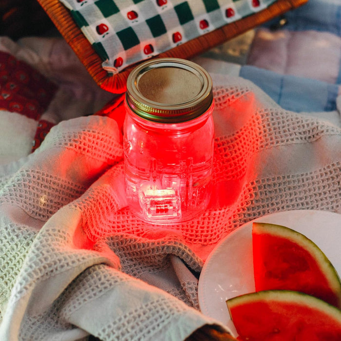 Red light-up cube glowing inside a water-filled mason jar on a picnic blanket next to watermelon slices.