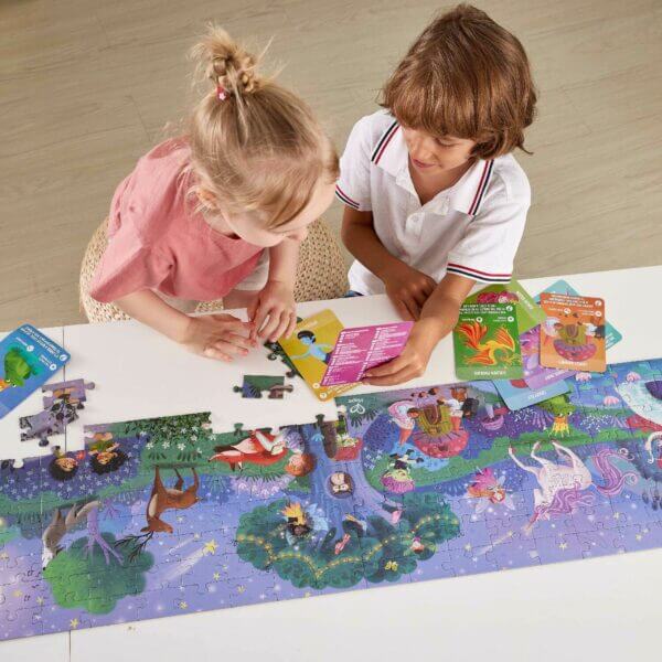 Two children assemble a long, colorful magical forest jigsaw puzzle with character cards on a white table.