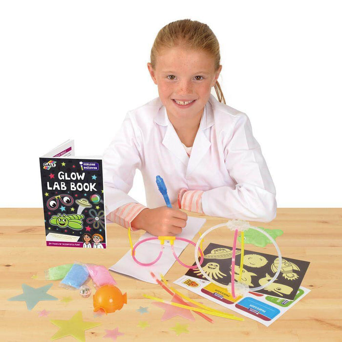 Child in a lab coat with a Glow Lab book, glow stick glasses, and neon accessories on a wooden table.