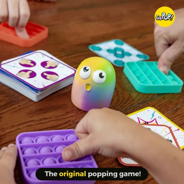 Hands pressing colorful bubble poppers around a rainbow character with big eyes and game cards on a wooden table.