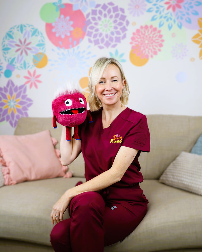 Woman holds a fuzzy pink monster puppet with white tufted hair, big eyes, and a wide toothy mouth.