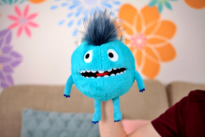 Round bright blue fuzzy monster puppet with large eyes, a wide toothy grin, and spiky gray hair held on a hand.