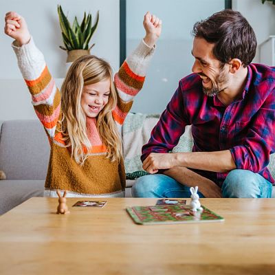 A girl cheers while playing a board game with rabbit figurines and cards as a smiling man watches.