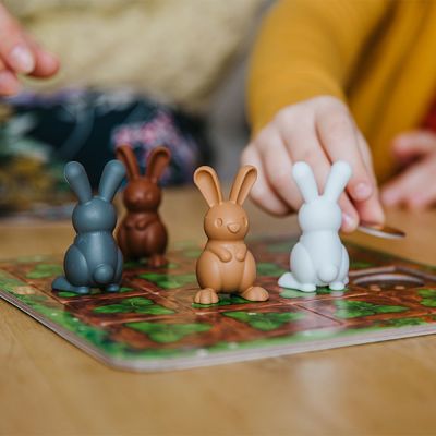 Four plastic bunny game pieces in grey, brown, tan, and white on a garden-themed game board with hands playing.