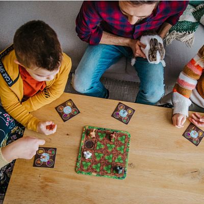 Family playing a garden board game with rabbit figurines and vegetable cards on a light wood coffee table.