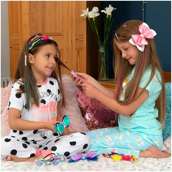 Two girls in pajamas play with colorful hair bows, headbands, and a hair styling tool on a bed.