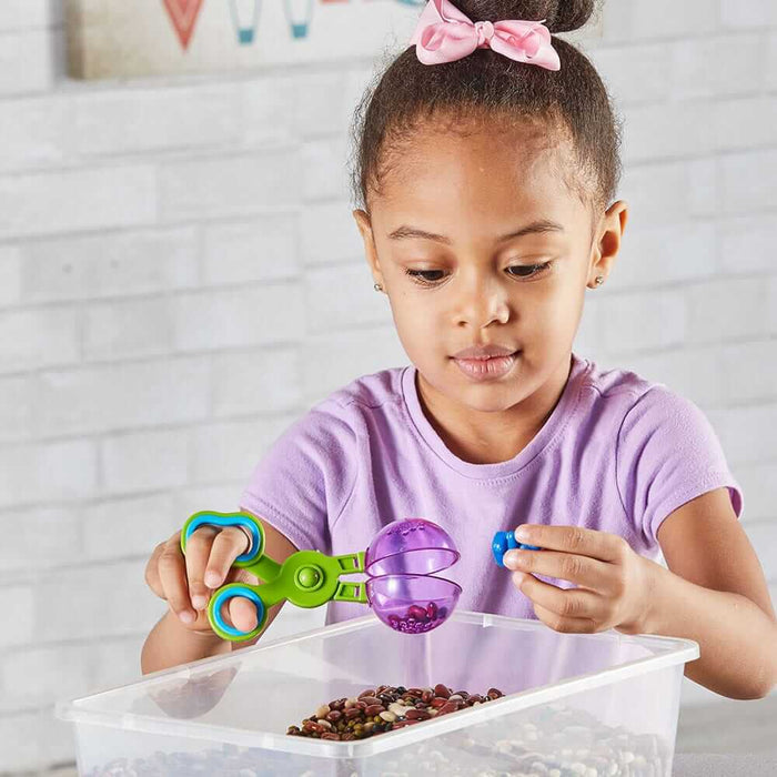 A girl uses a green and blue toy scooper with a purple spherical end to pick up dried beans from a clear bin.