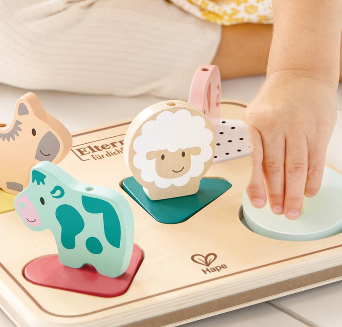 Child's hand placing chunky wooden animal pieces, like a green cow and white sheep, into a wooden puzzle board.